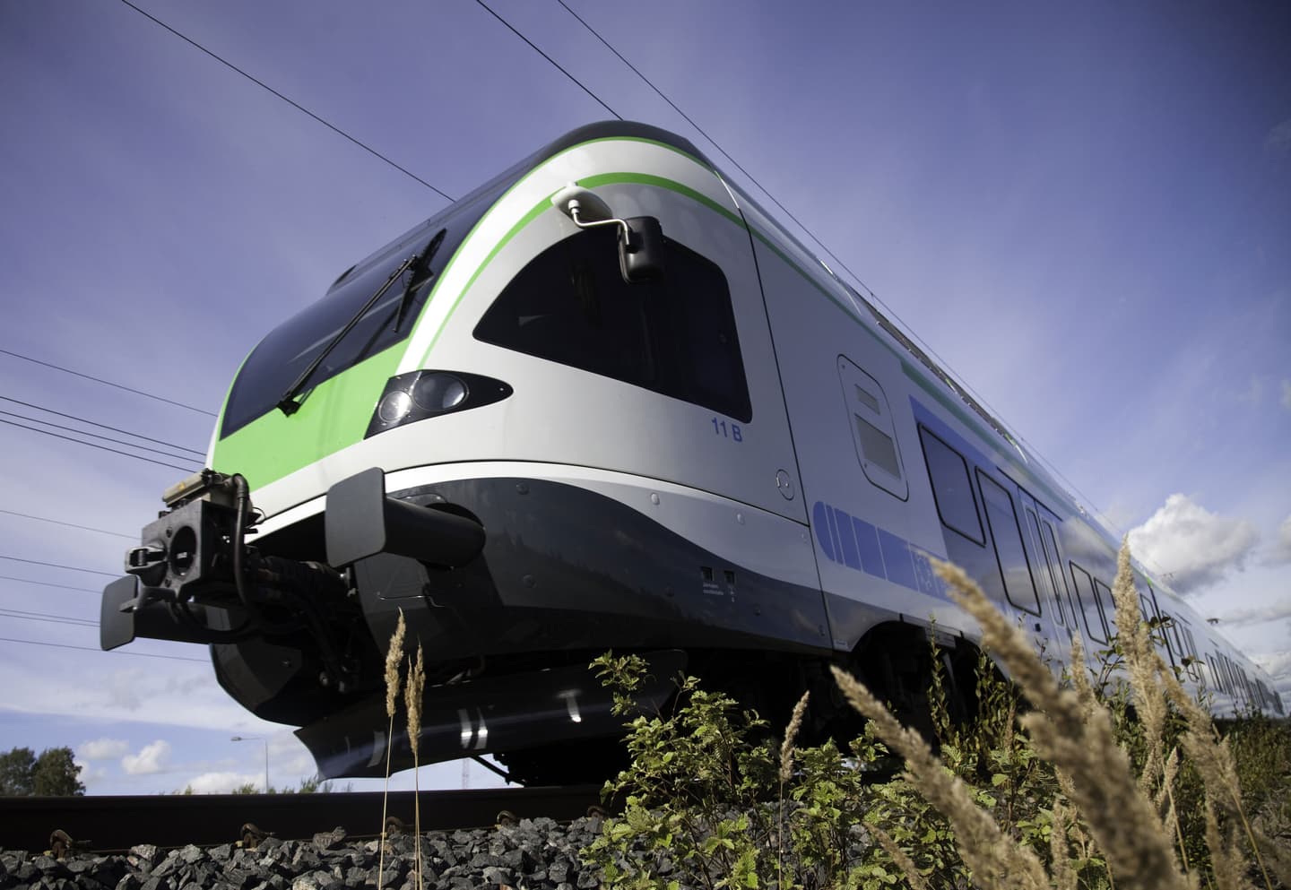 Modern passenger train with white and green livery travelling on tracks against blue sky, viewed from low angle.