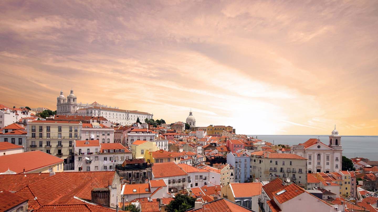Lisbon cityscape with orange terracotta rooftops, historic buildings and cathedral under a golden sunset sky.