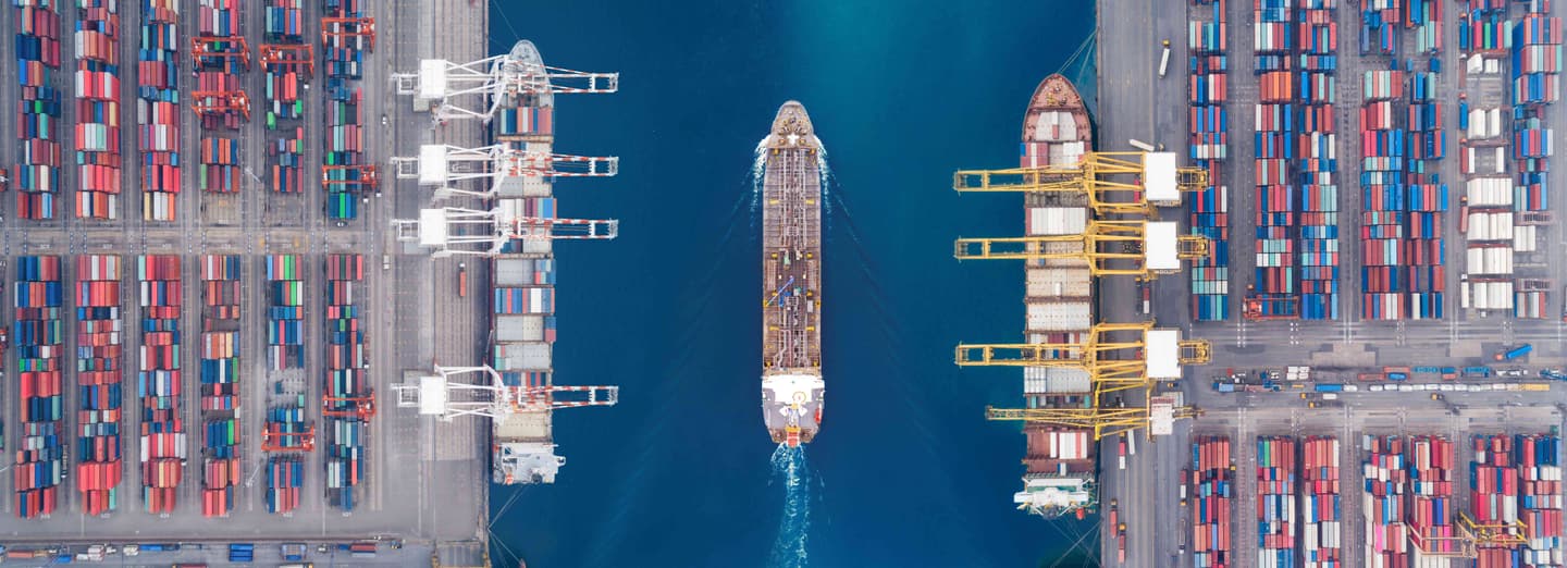 Aerial view of a busy shipping port with cargo vessels, colourful shipping containers, and loading cranes beside deep blue water.