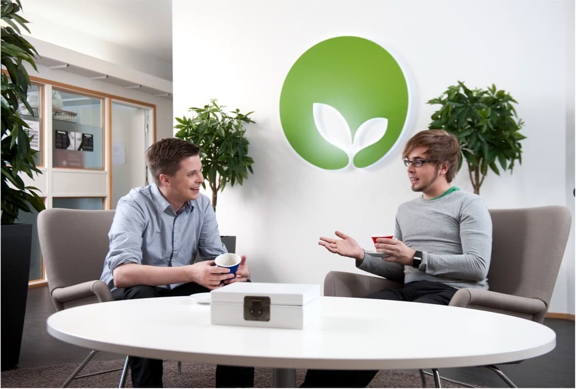 Two people conversing in modern office with green eco-friendly logo on wall and indoor plants surrounding them.