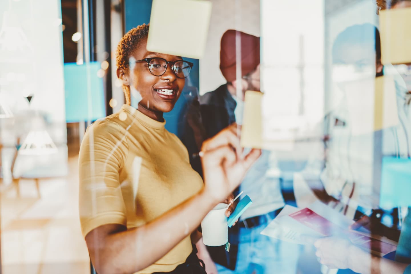 Person in yellow top and glasses smiling while placing sticky notes on a glass wall during a collaborative session.