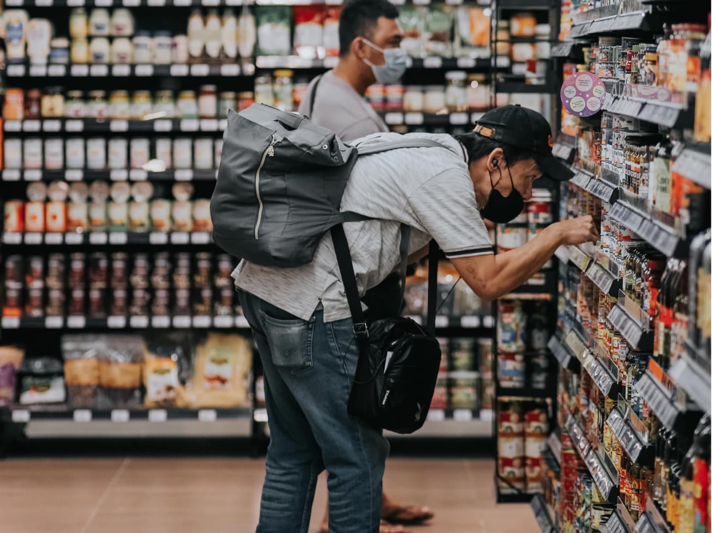 Shoppers wearing face masks browse grocery store shelves, one with a grey backpack examining products closely.
