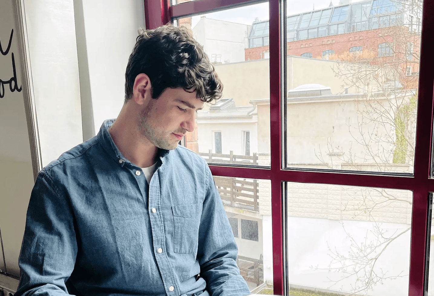 Person in blue denim shirt looking down thoughtfully by large window with urban buildings visible outside.