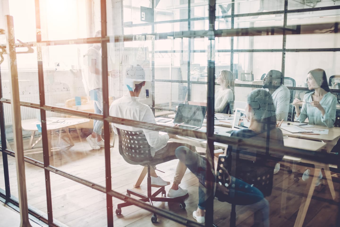 Modern office meeting viewed through glass partition, with diverse professionals collaborating around laptops in sunlit space.
