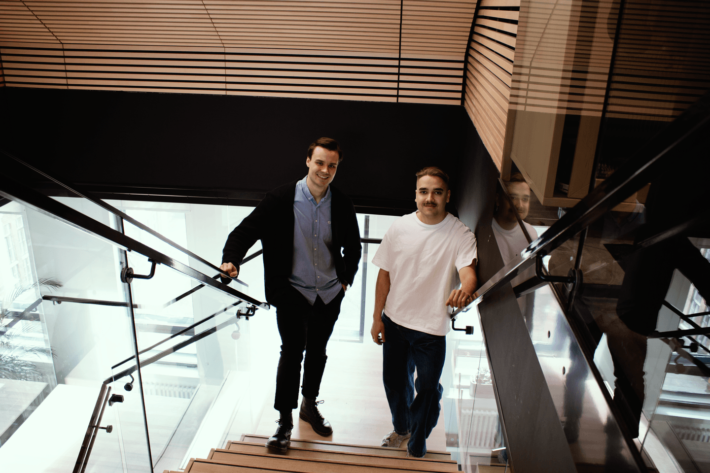 Two young men standing on a modern staircase with glass railings in a contemporary building with wooden details.
