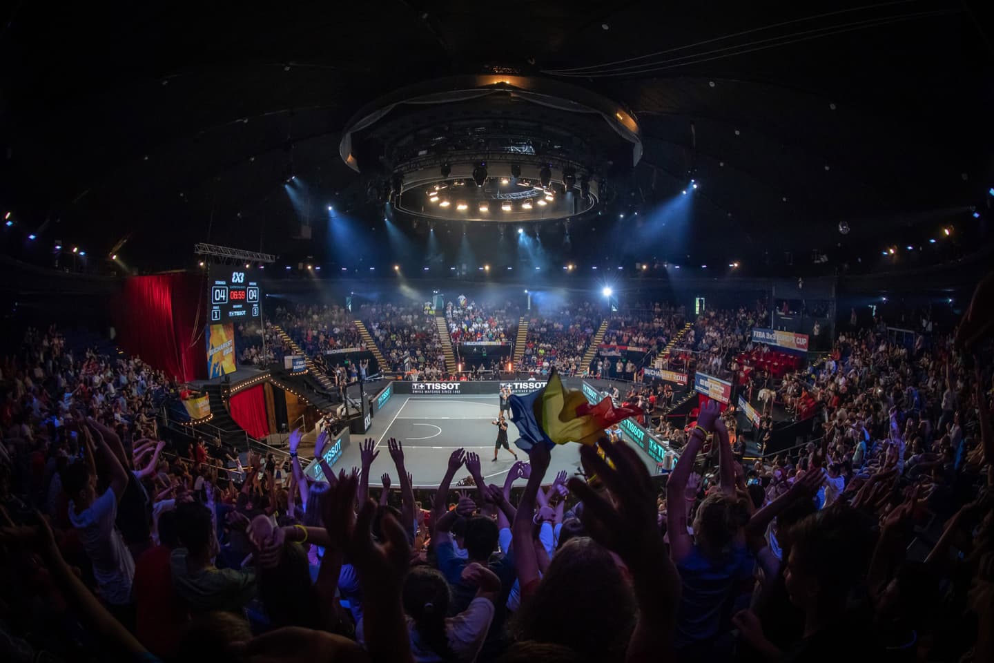 Indoor basketball arena with enthusiastic crowd watching a FIBA 3x3 tournament, dramatic lighting and scoreboard visible.