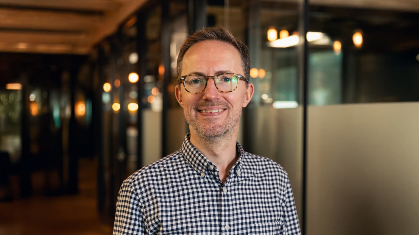 Matthew Billson of Piclo Energy in a checkered shirt, smiling ina modern office space with warm lighting in the background.