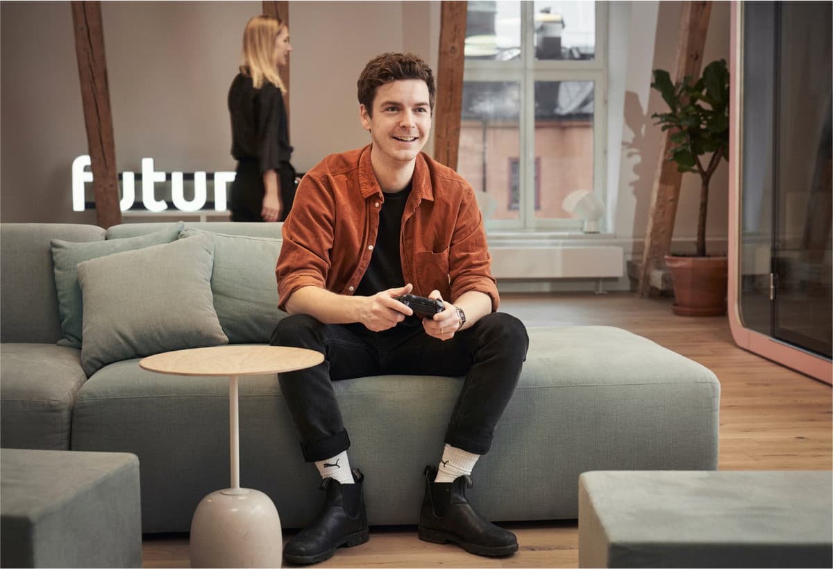 Person in rust-coloured shirt playing video games on grey sofa in modern office with 'futur' sign visible.