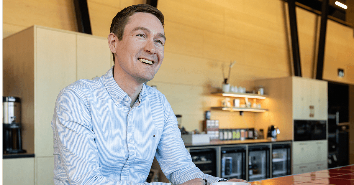 A smiling person in a blue striped shirt sitting in a modern kitchen with wooden walls and minimalist cabinetry.