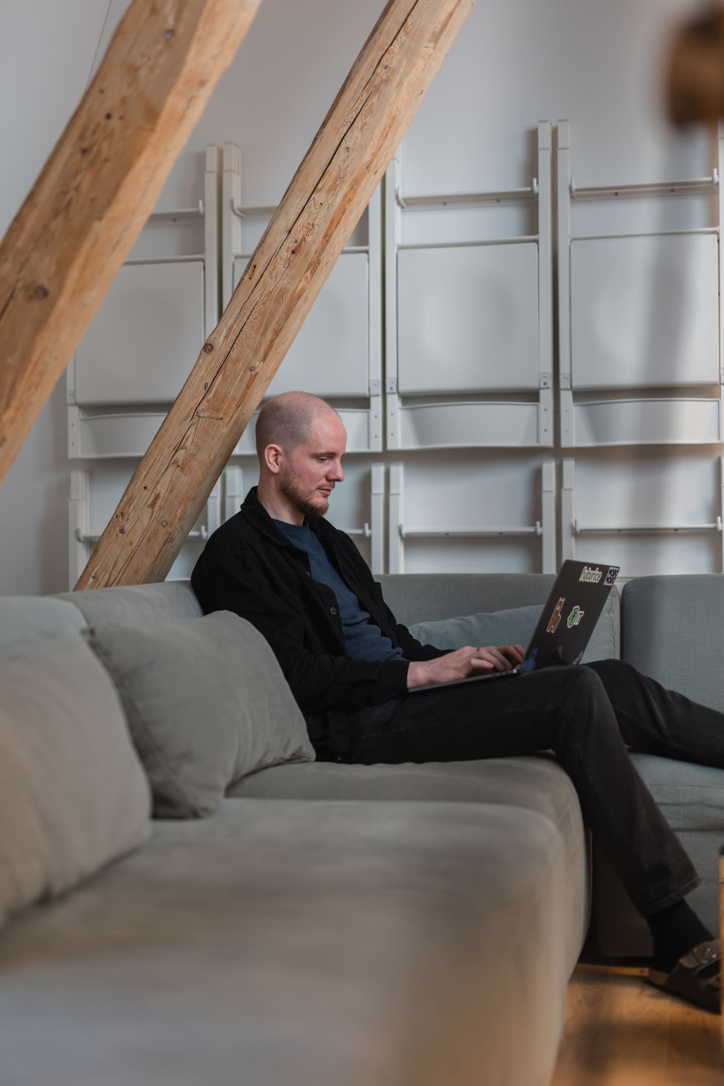 Person working on laptop on grey sofa in modern room with exposed wooden beams and white shelving.