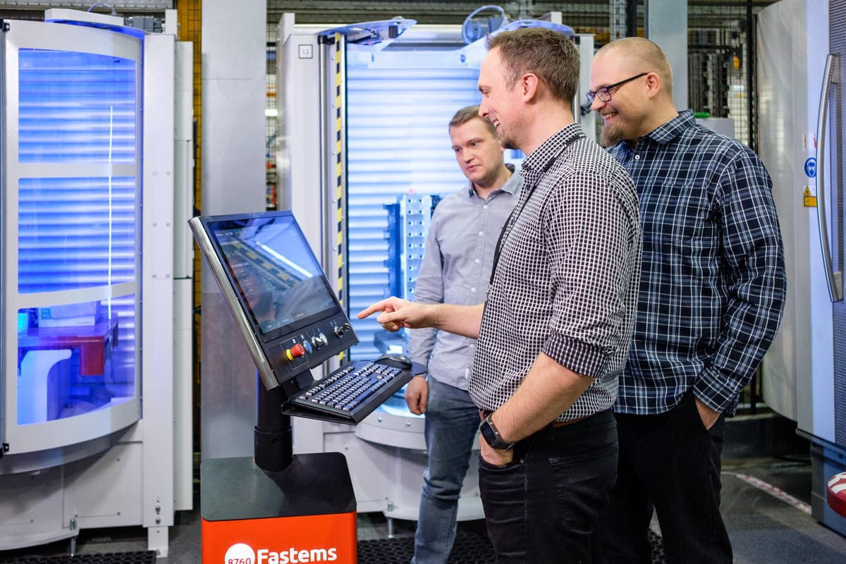 Three colleagues in a manufacturing facility operating a control panel with blue-lit industrial equipment in the background.