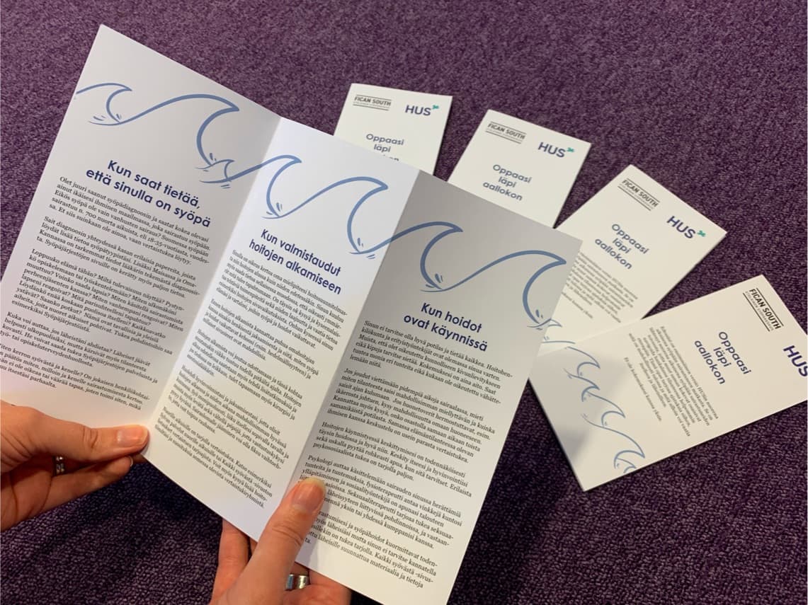 Hands holding Finnish healthcare brochures with blue wave designs on a purple carpet, HUS medical information pamphlets.