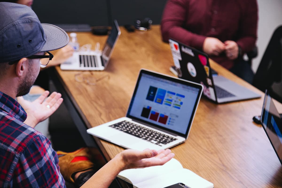 People working around a wooden table with laptops displaying design layouts, one person wearing a blue cap.