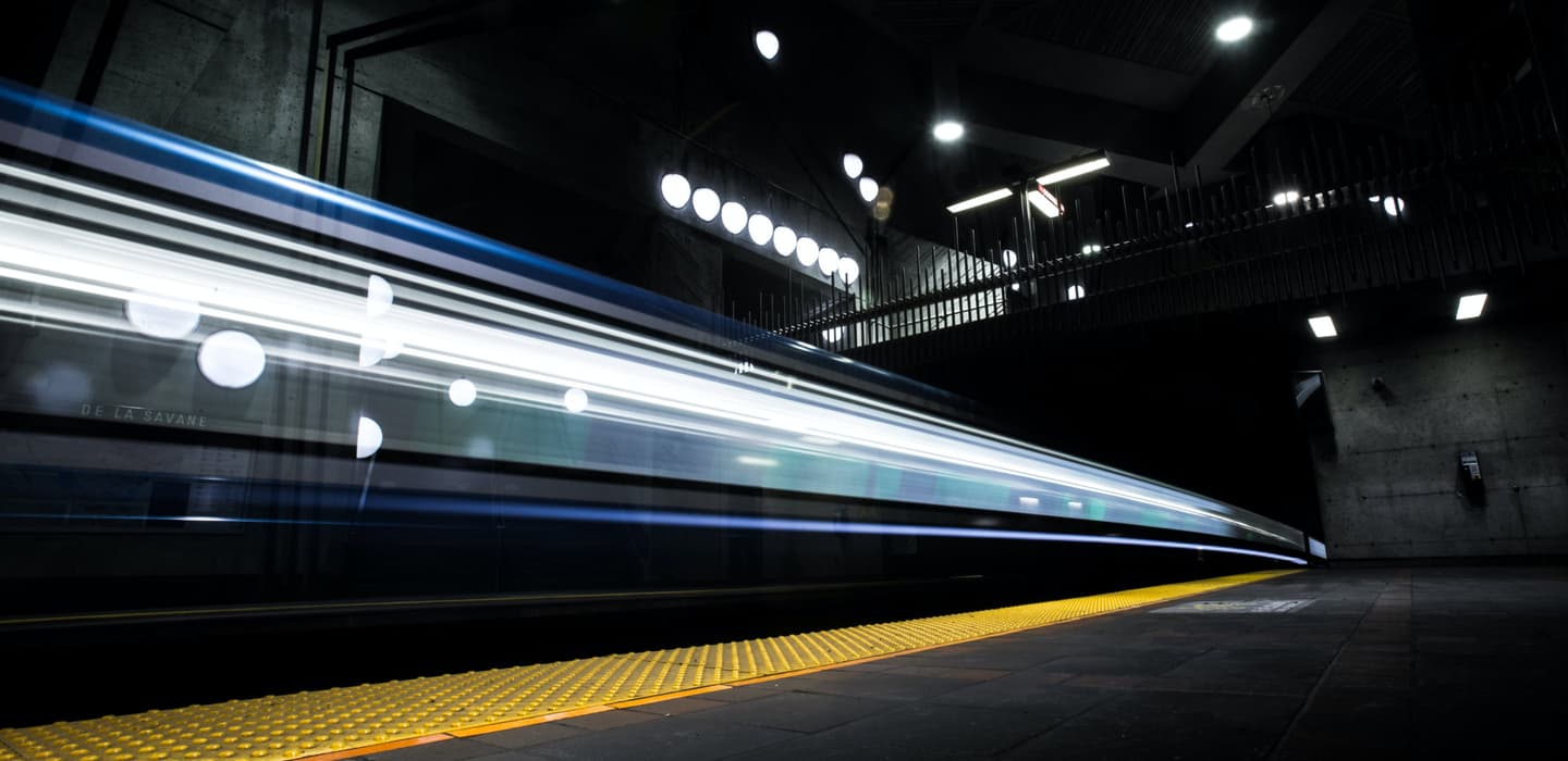 Blurred train speeding through dark subway station with yellow safety strip along platform edge and overhead lights.