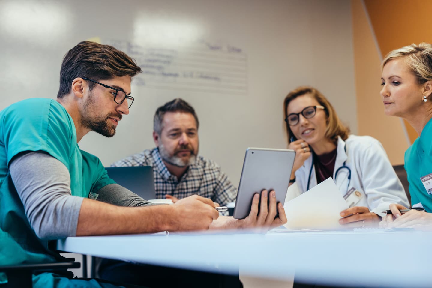 Medical team in scrubs and white coat reviewing information on a tablet during a consultation meeting.