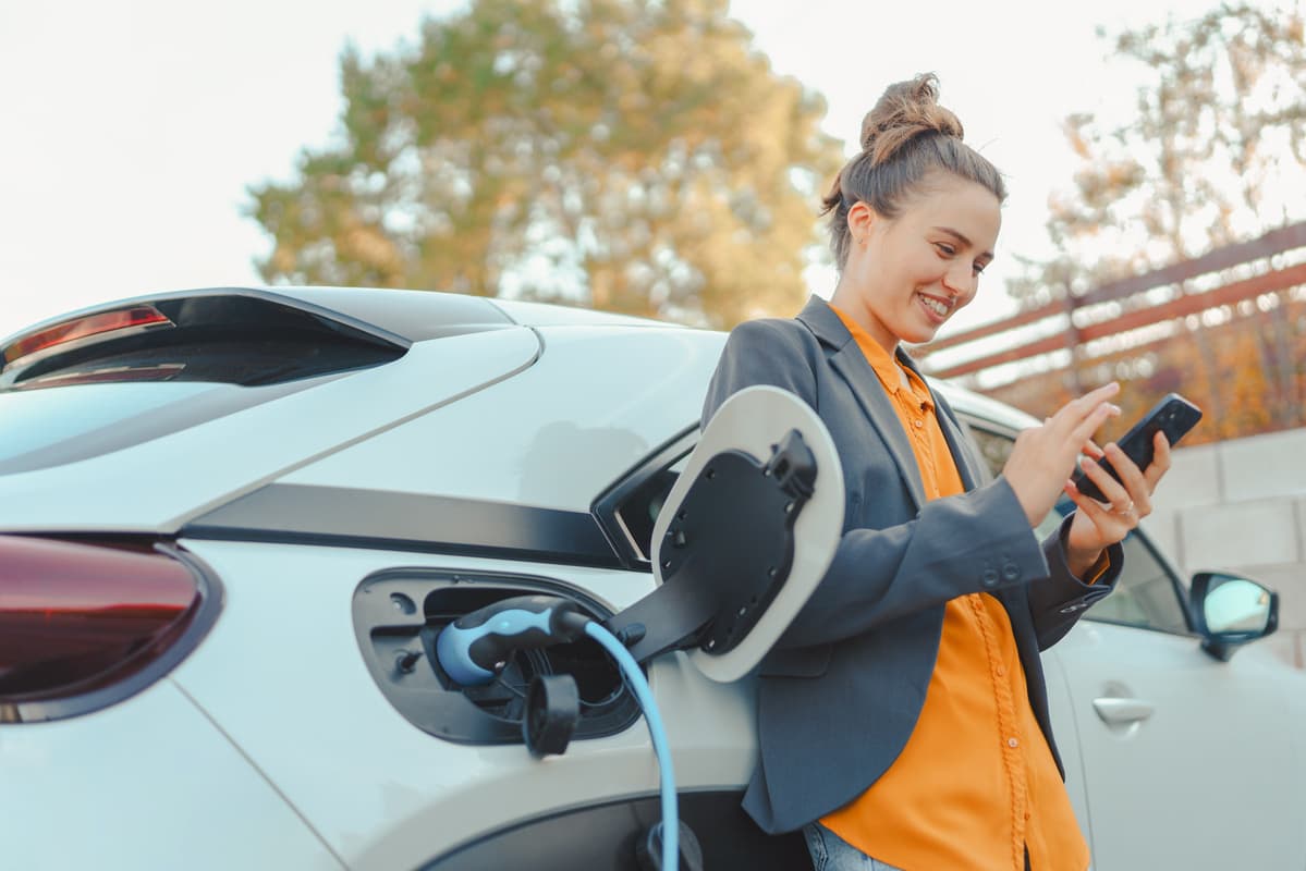 Person in grey blazer and orange shirt using smartphone while charging electric vehicle outdoors in autumn setting.