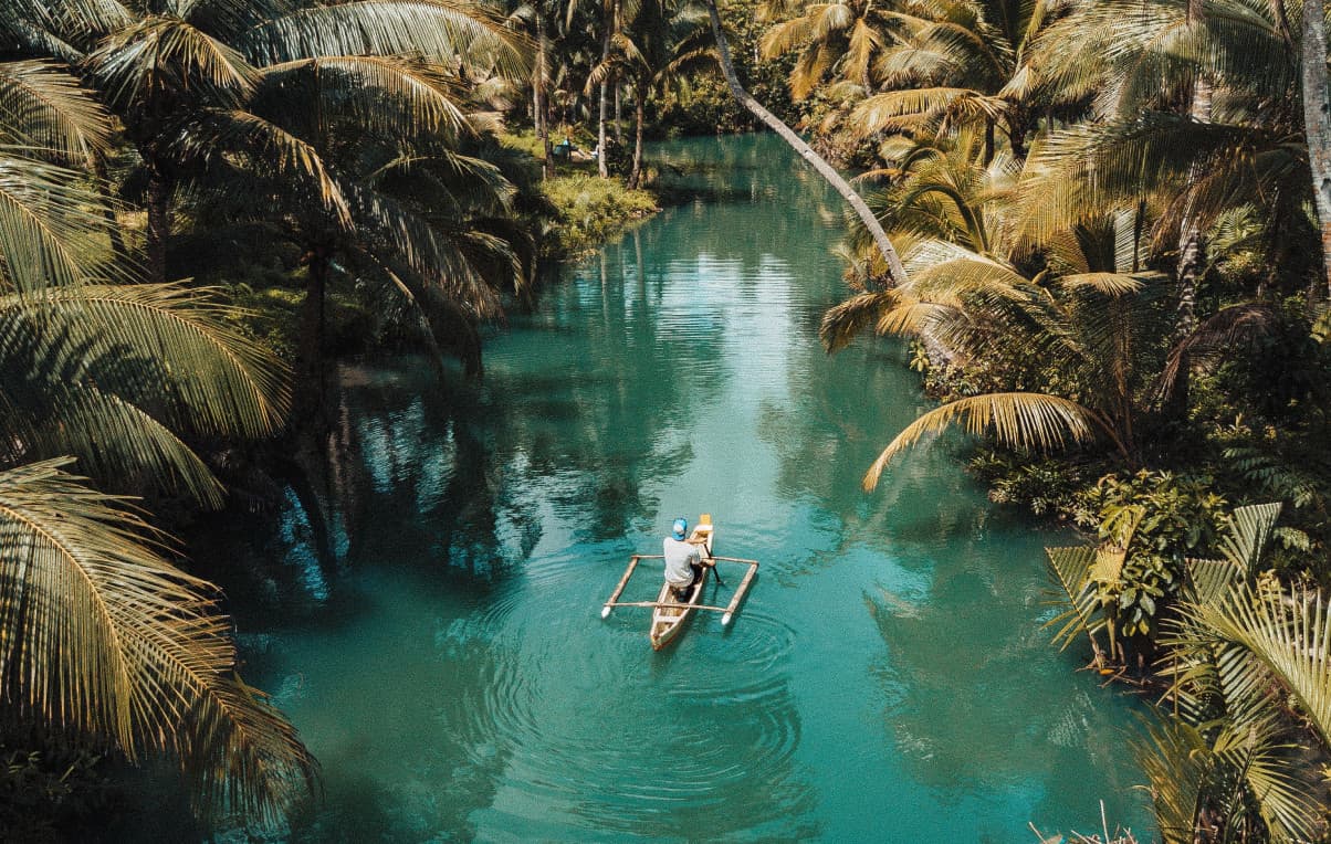 Person paddling a small outrigger canoe through turquoise water surrounded by lush palm trees in a tropical setting.