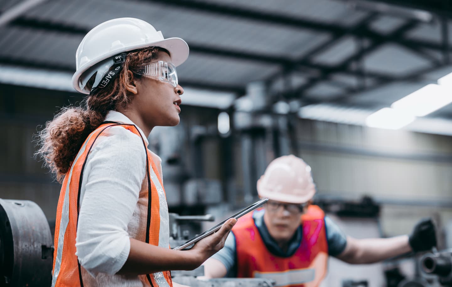 Construction worker in white helmet and orange safety vest holding tablet in industrial setting with colleague nearby.