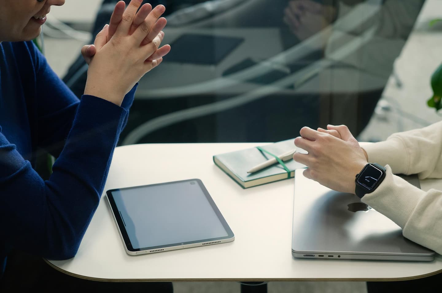 Two people in discussion at white table with tablet, laptop, notebook and pen. One wears blue, the other a smartwatch.