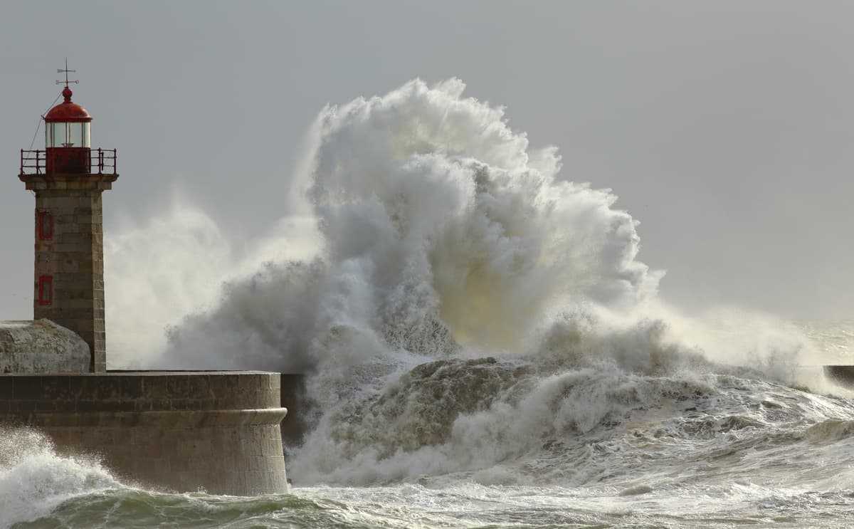 Massive wave crashes against harbour wall with red-topped lighthouse standing firm against stormy sea.