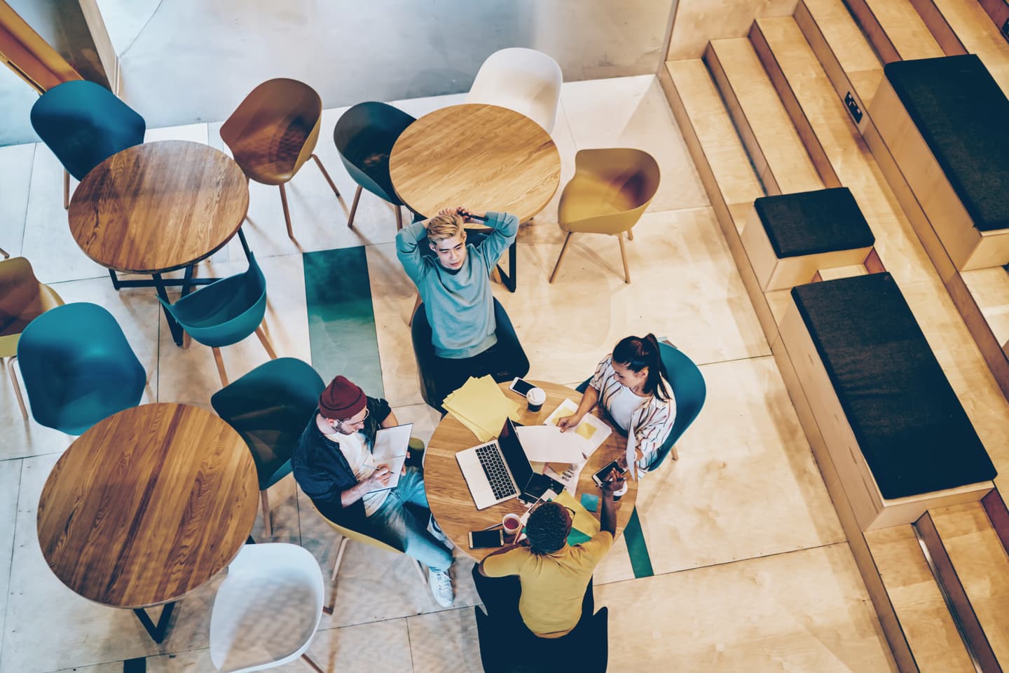 Overhead view of young people working around a wooden table in a modern café with colourful chairs and wooden steps.