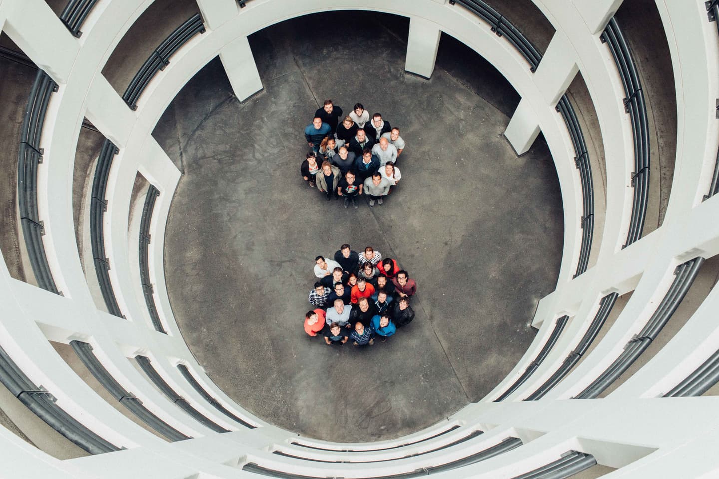Aerial view of two groups of people standing in circular formations on a concrete floor surrounded by curved white architecture.