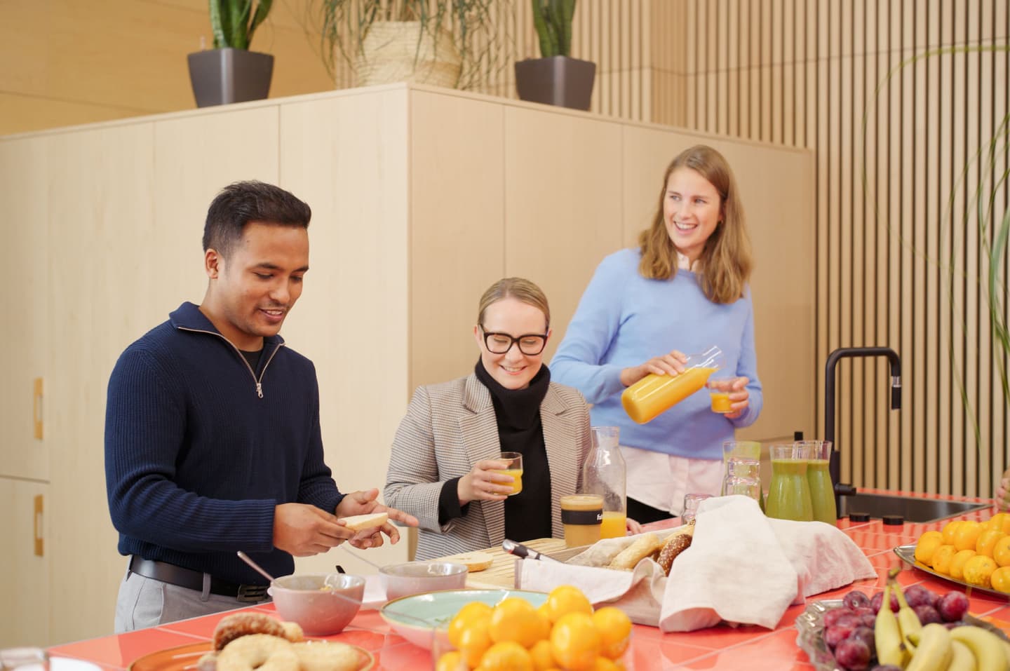 Three colleagues enjoying breakfast in a modern office kitchen with fresh juice, fruit and pastries on a coral-coloured table.