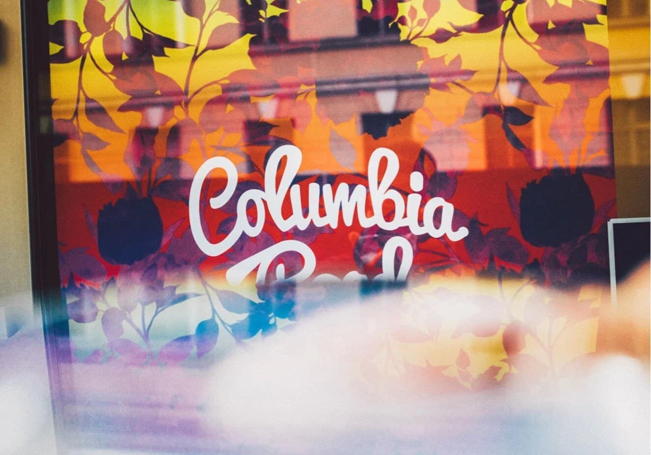 Columbia Road sign with floral patterns on colourful gradient background, viewed through glass.
