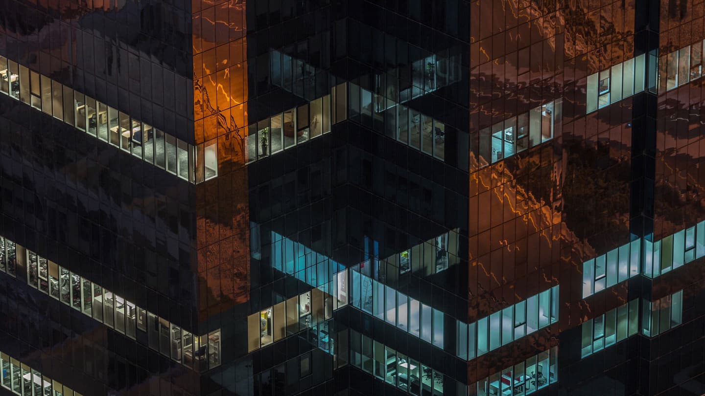 Modern office building at night with illuminated windows and copper-coloured glass reflections on its facade.