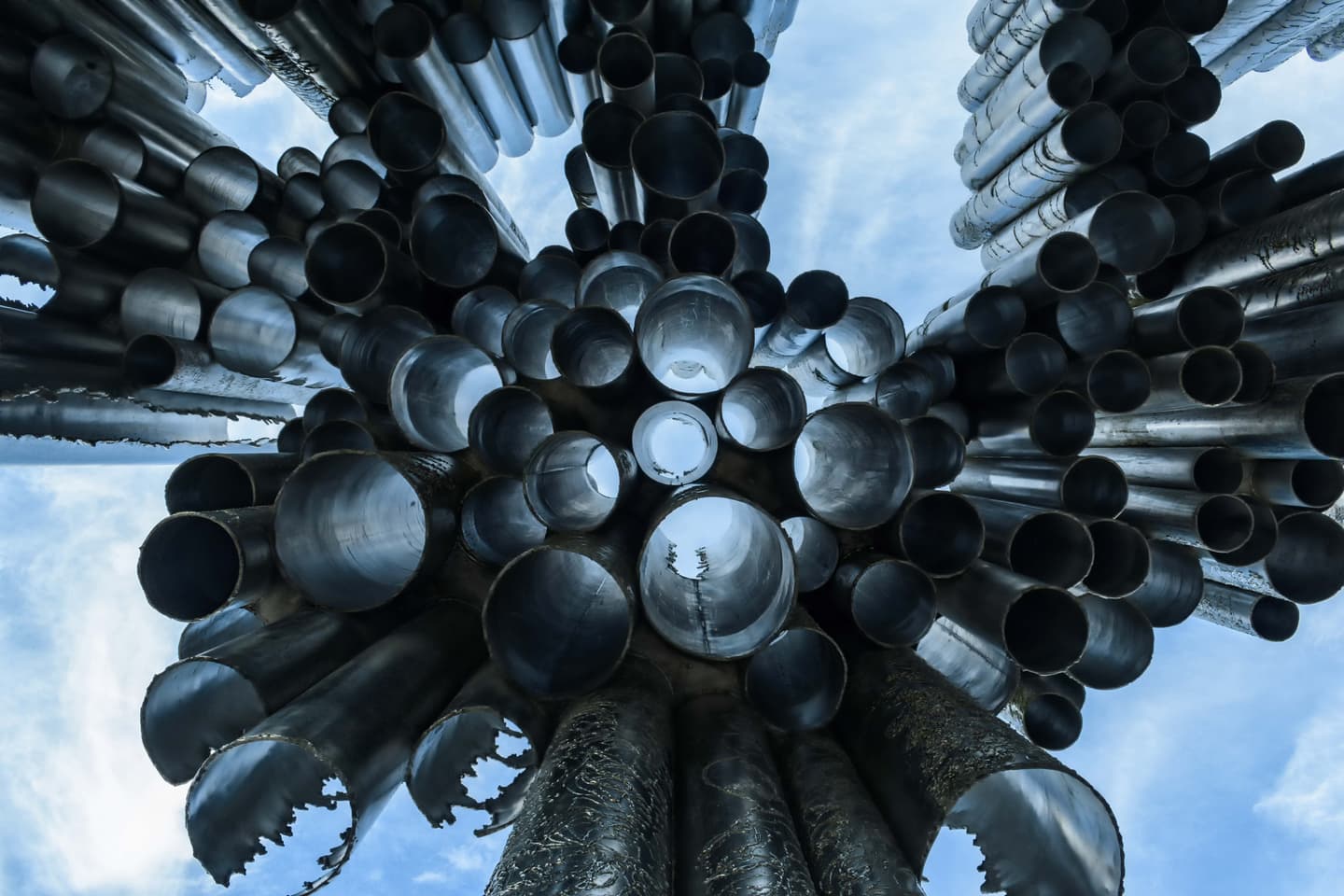 Looking up at a sculptural installation of metal pipes arranged in a circular pattern against a blue sky.