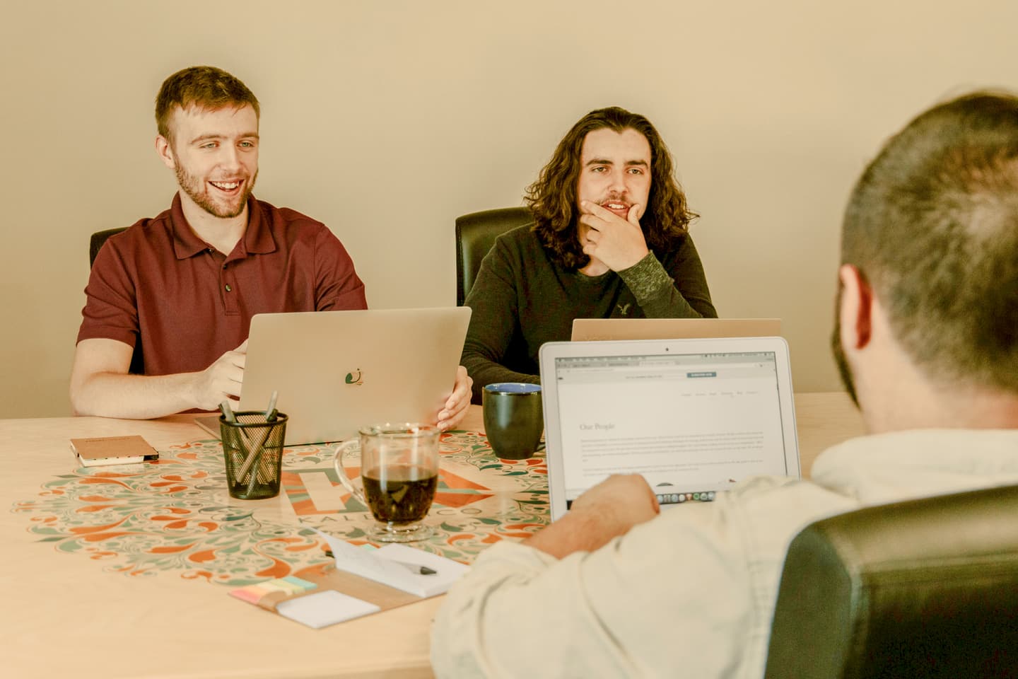 Three people in a meeting with laptops on a patterned table with coffee mugs and office supplies.