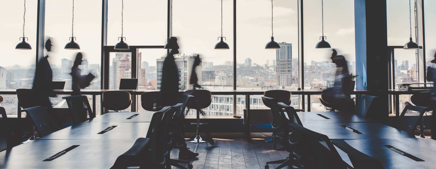 Blurred figures moving in a modern office space with large windows overlooking a city skyline and pendant lighting.