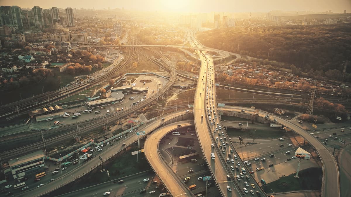 Aerial view of a city motorway interchange at sunset, with winding roads, traffic and high-rise buildings in golden light.
