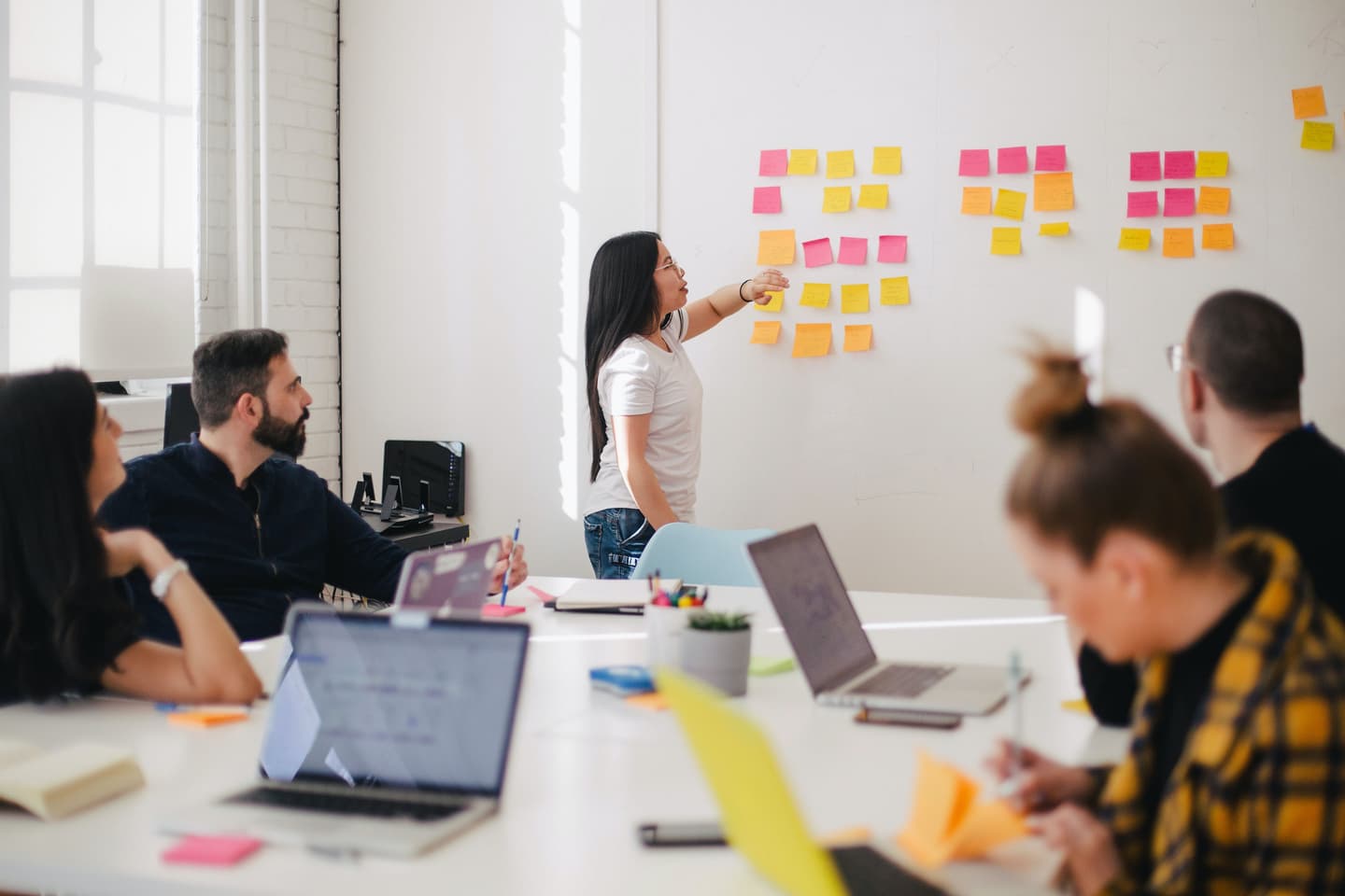 Woman pointing at colourful sticky notes on wall during team brainstorming session in bright office space.