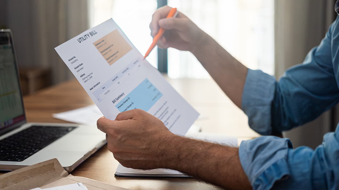 Person in blue shirt reviewing a utility bill with orange pen, laptop visible on wooden desk in bright room.
