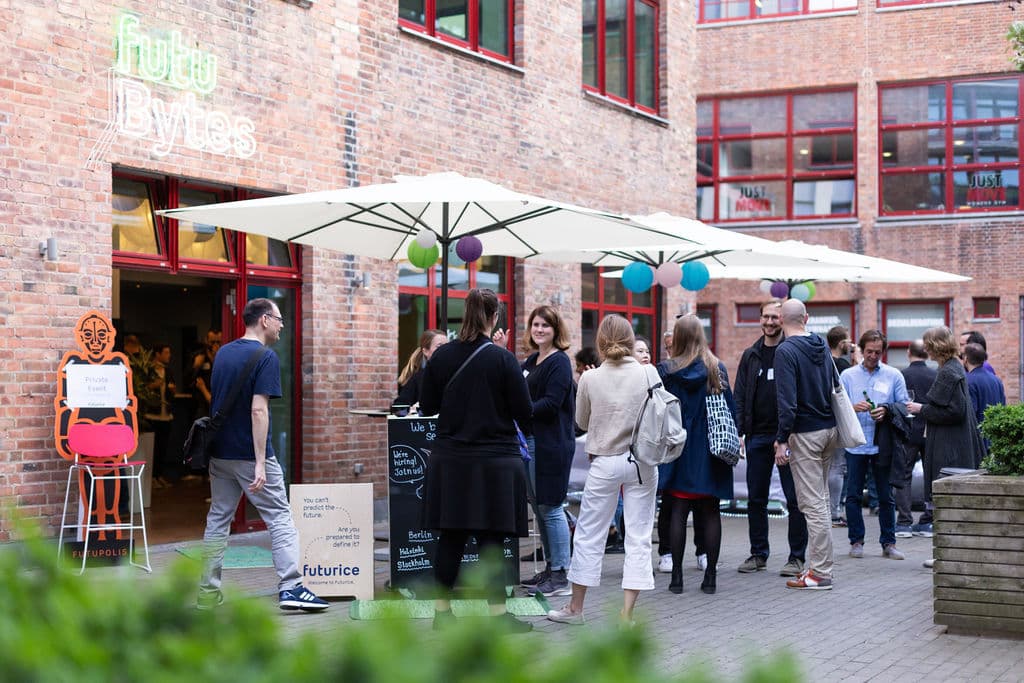 People gathering outside a brick building with "hub.berlin" sign, under white umbrellas with colourful balloons.