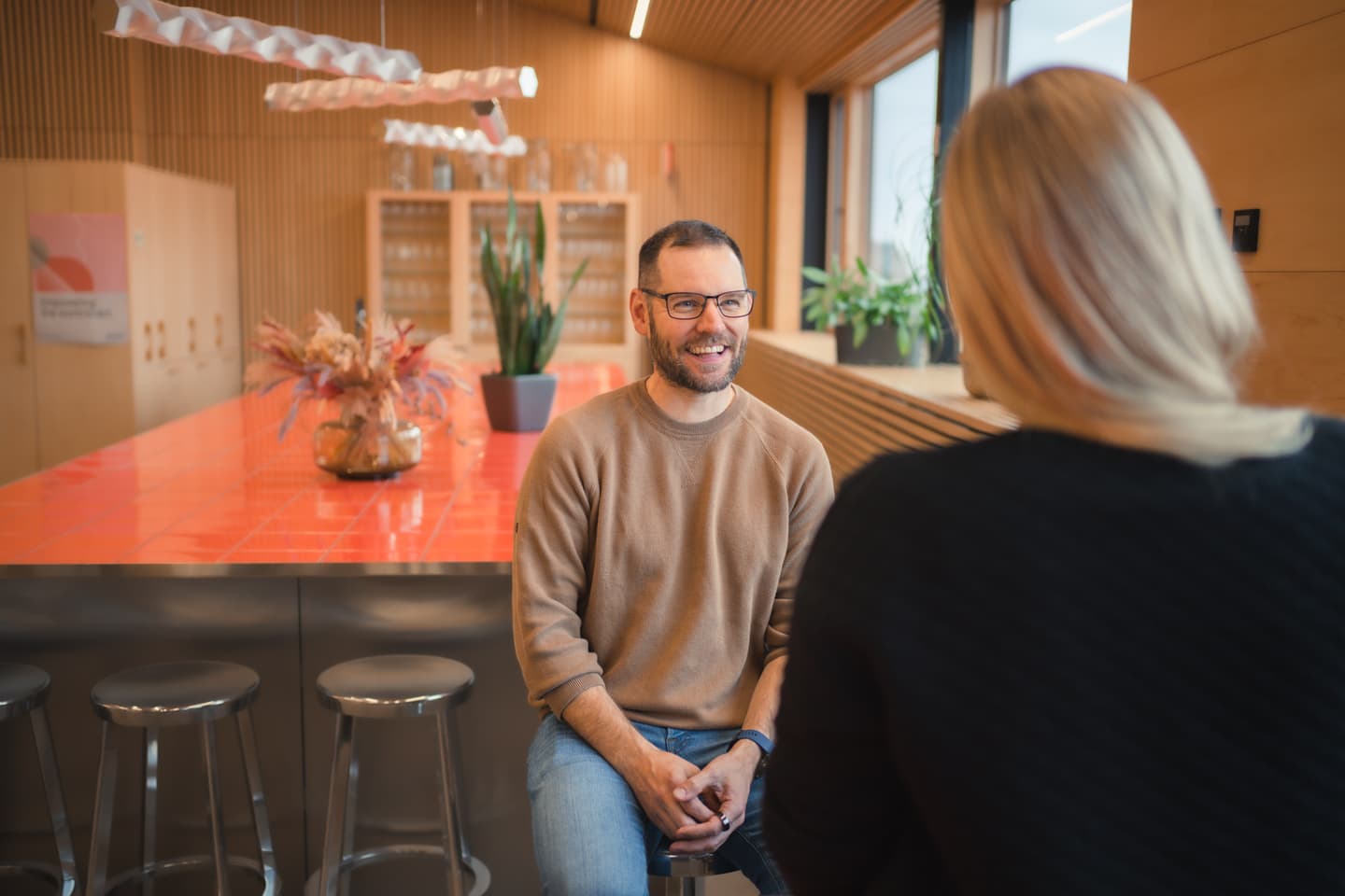 Two people conversing in a modern office space with wooden walls, orange countertop, and potted plants.